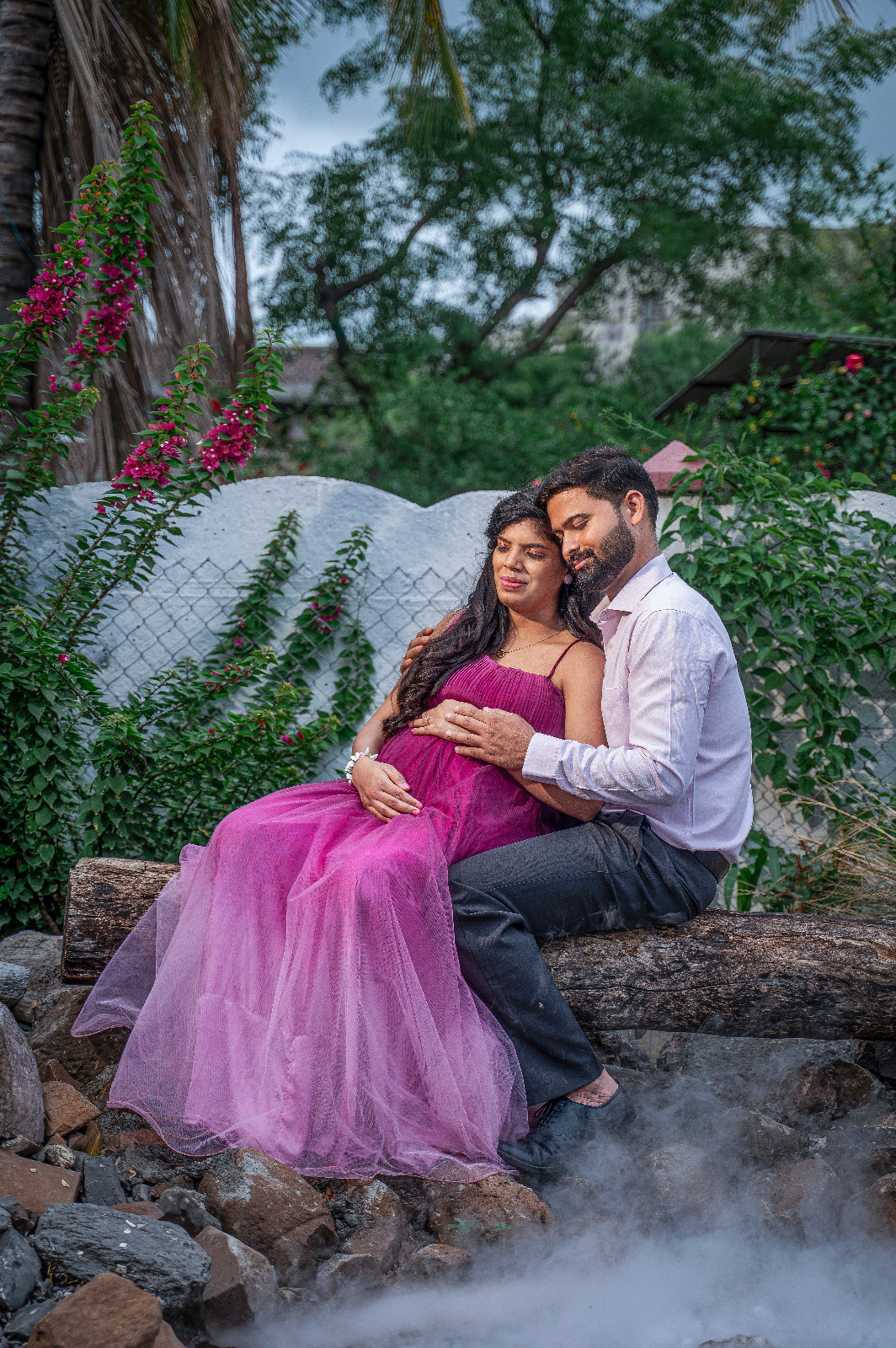 Expectant couple seated together outdoors with the mother wearing a flowing magenta gown