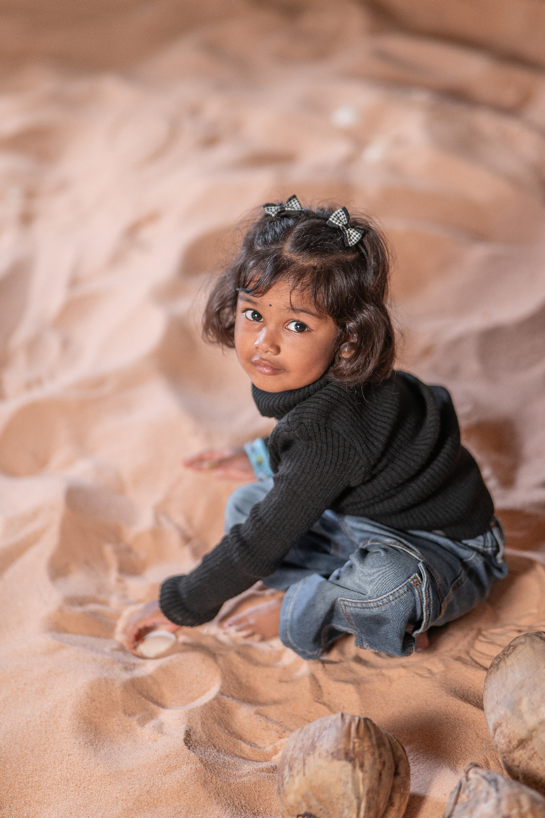 Young girl in a black sweater and blue jeans seated in sand among coconuts and looking toward the camera