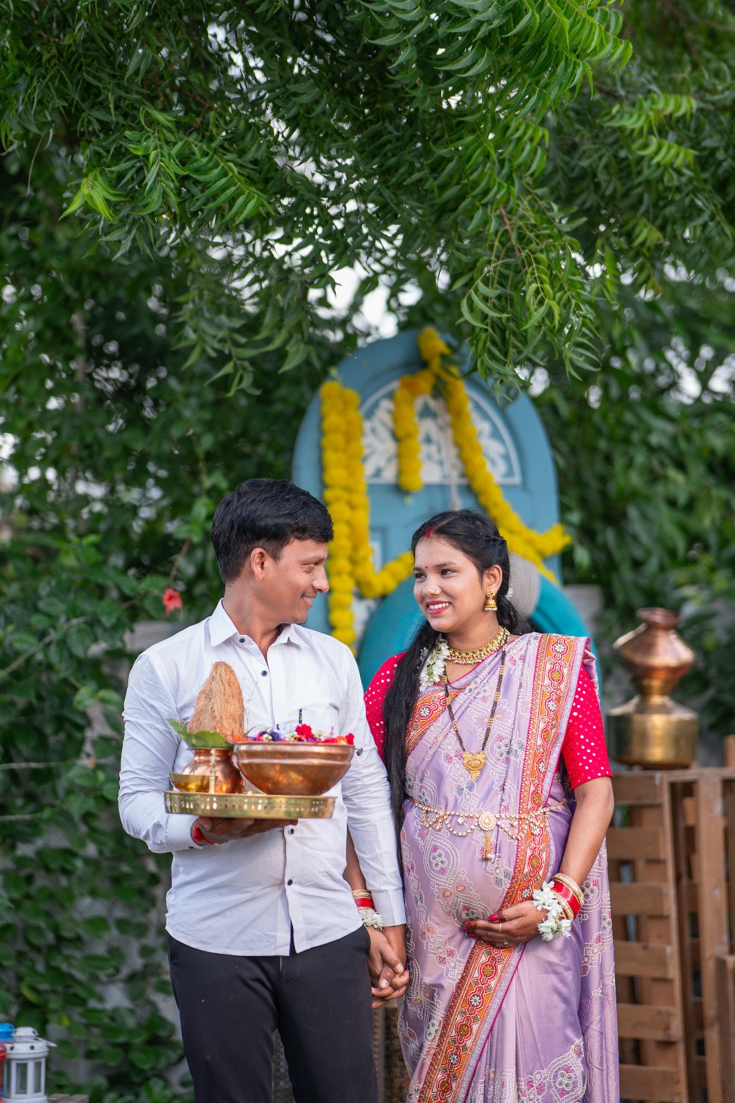 Expectant couple in traditional attire holding a ceremonial tray beneath a leafy neem tree