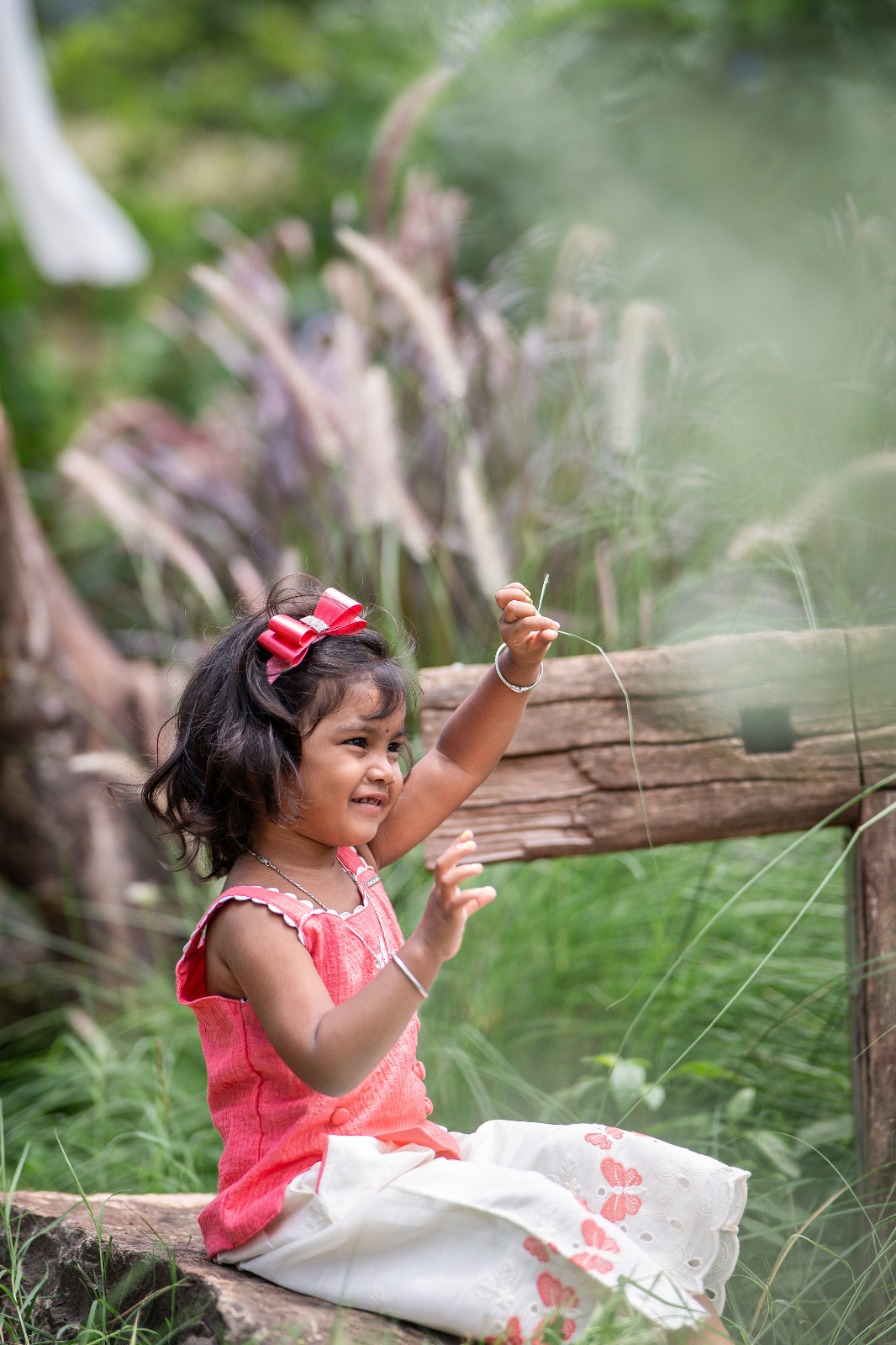 Young girl in a coral top and floral skirt playing with long grass beside a rustic wooden fence