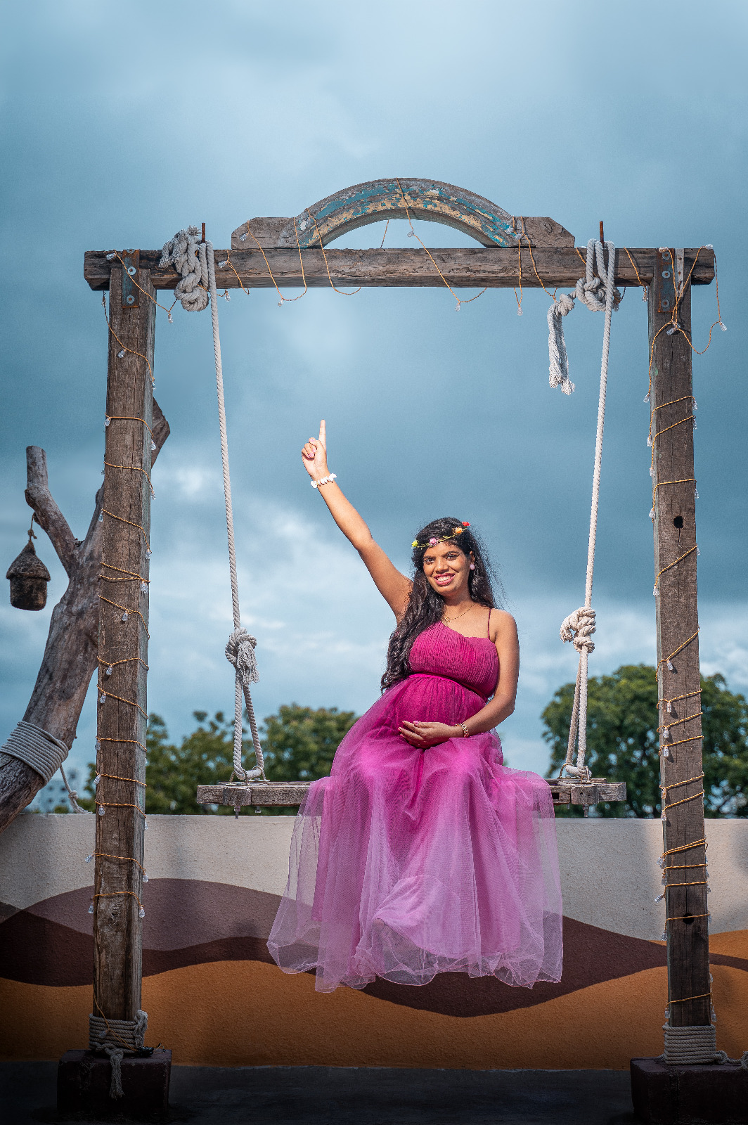 Smiling expectant mother in a magenta gown posing on a rustic outdoor swing