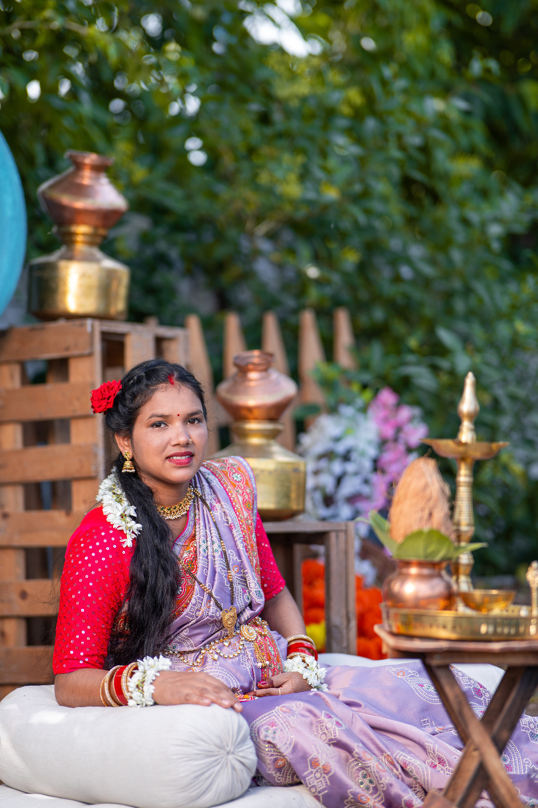 Expectant mother in a lavender saree seated beside traditional brass vessels in a garden setting