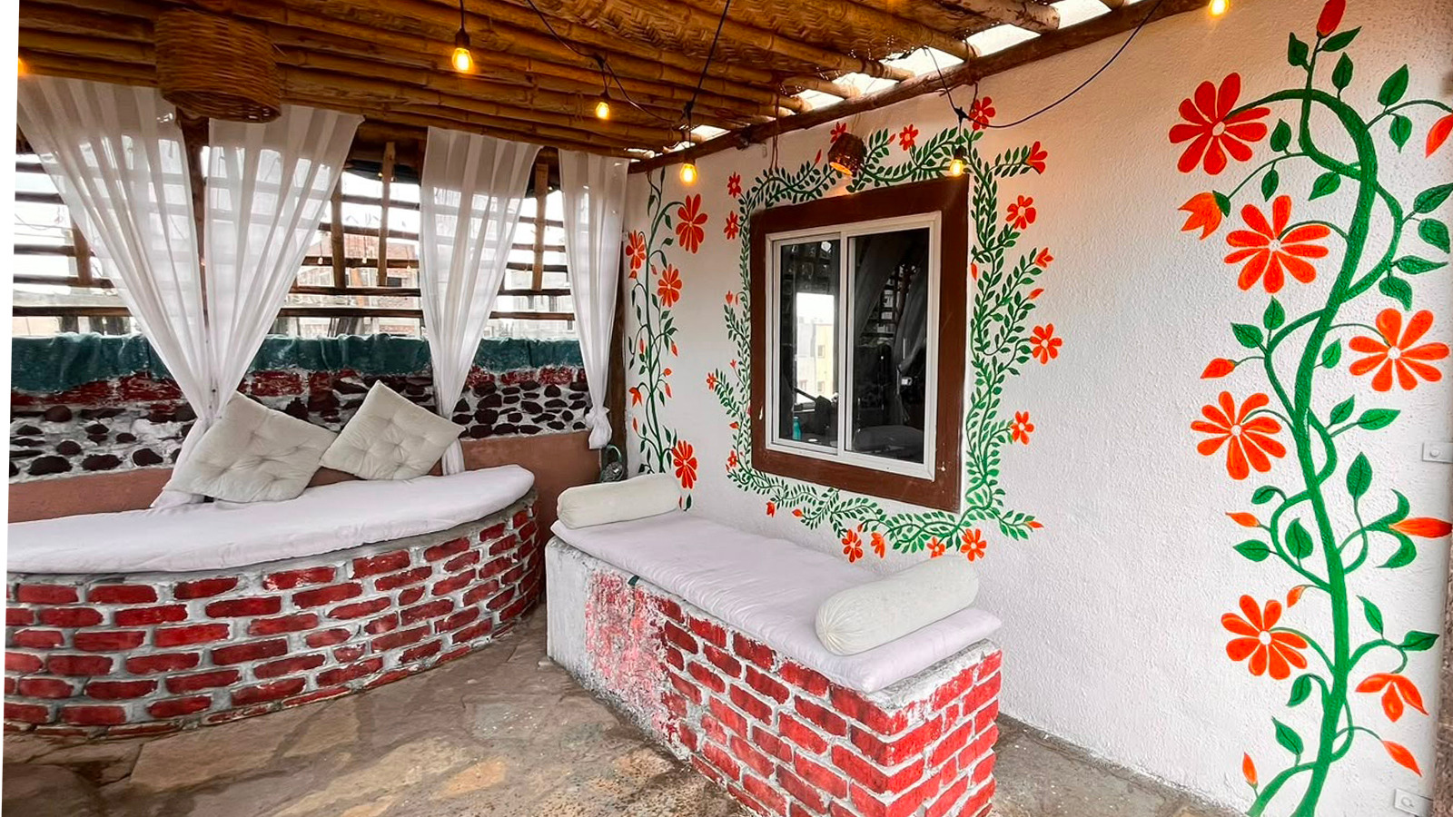 Covered studio lounge with red brick seating, white cushions, floral folk art and warm lights