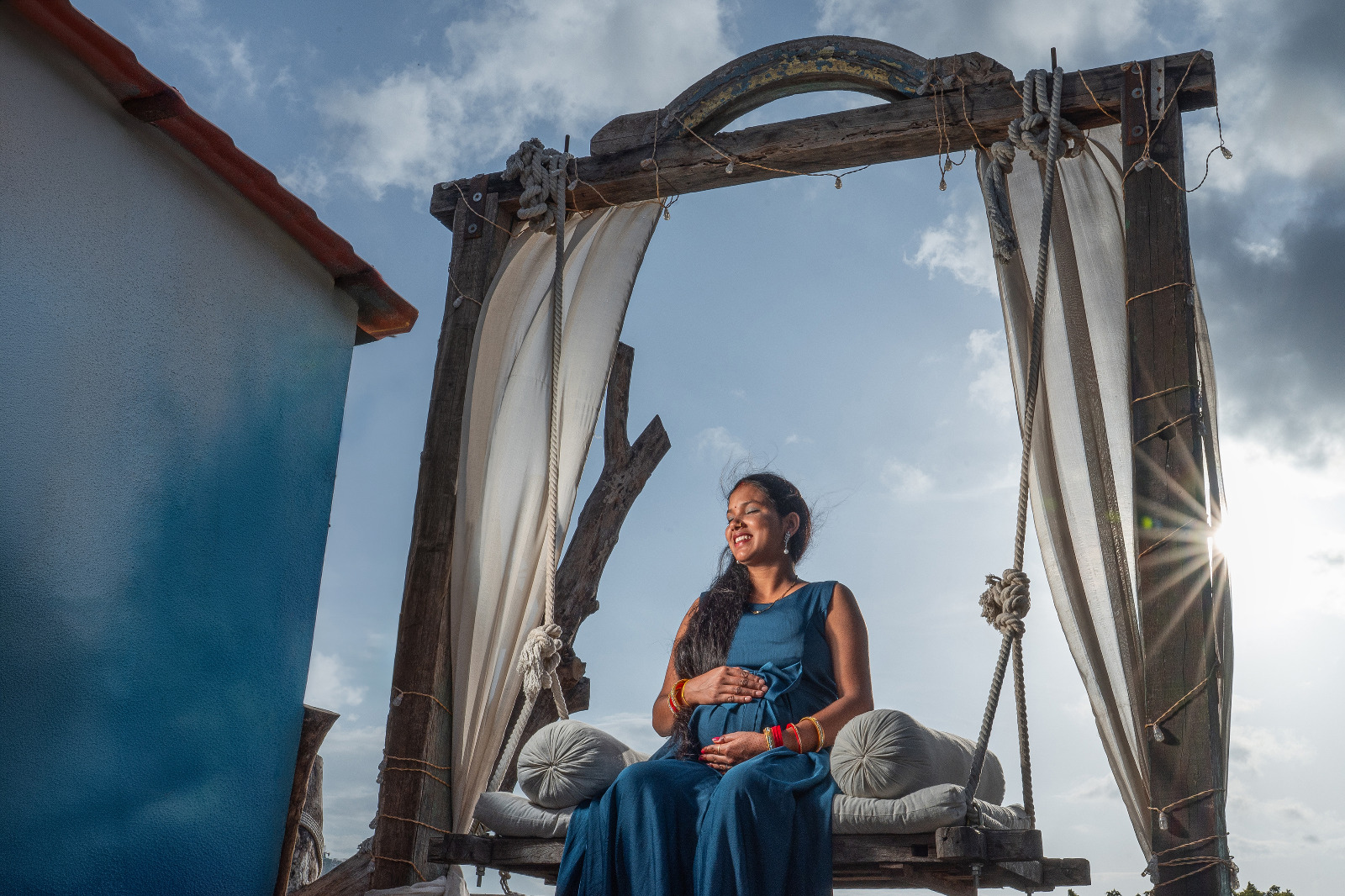 Expectant mother in a blue gown seated on a rustic outdoor swing with sunlight behind her