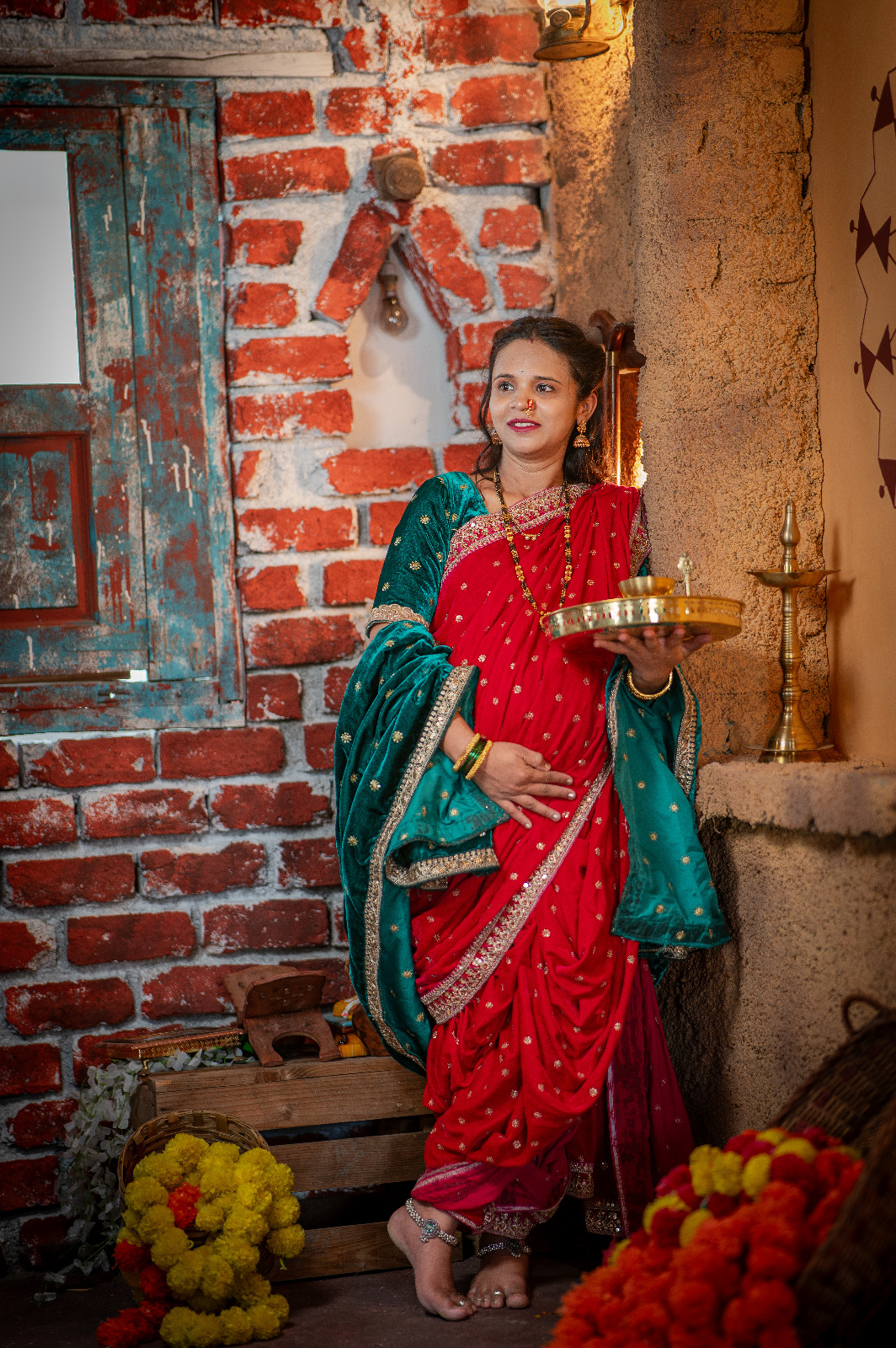 Expectant mother in a red and green saree holding a ceremonial brass tray beside a rustic doorway